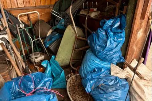 Vollgestellter Geräteschuppen mit Schubkarre, Gartengeräten und gefüllten Müllsäcken.